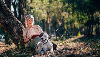 Older woman relaxes in forest with book and dog.